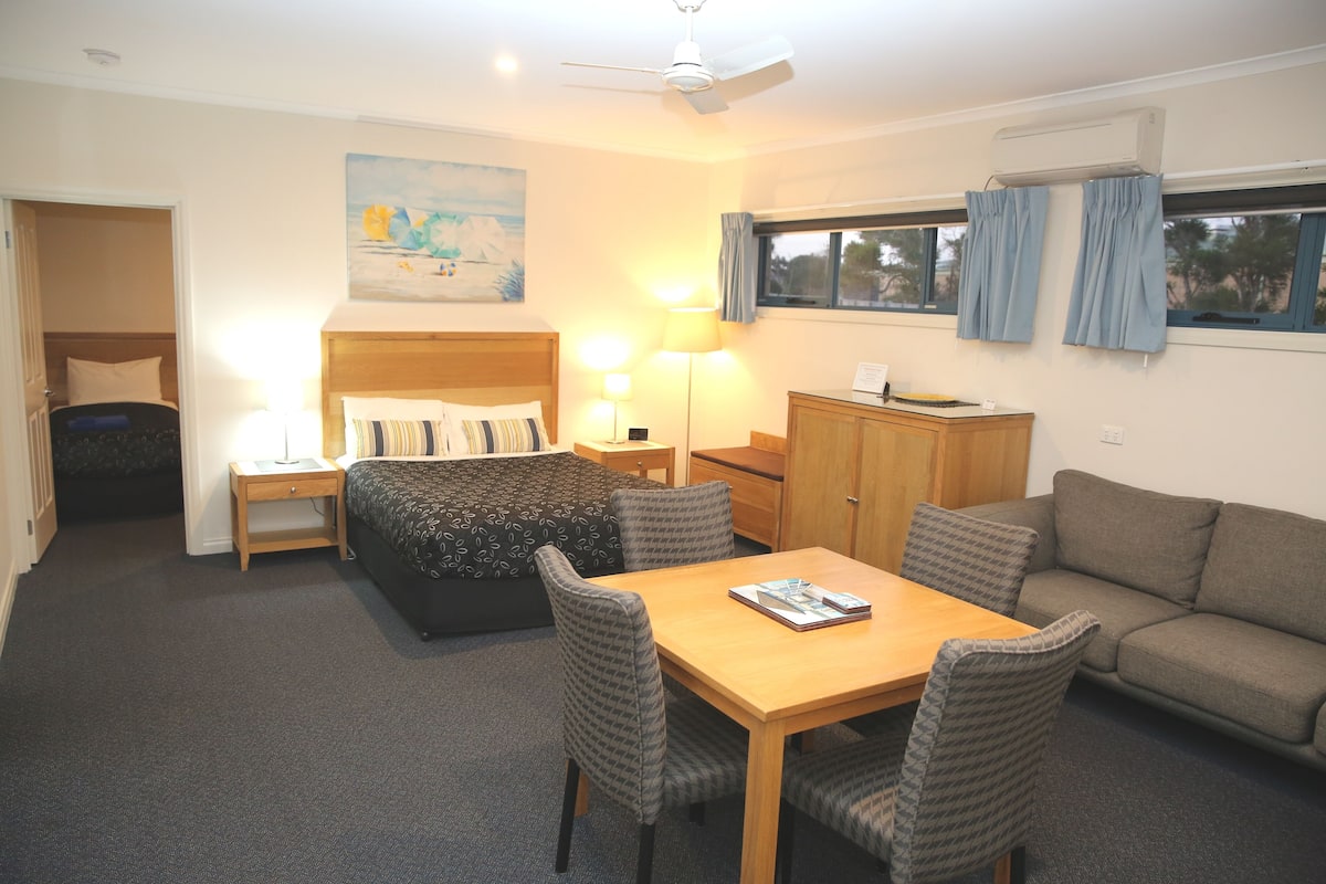 A hotel room with a bed, a sofa, a dining table with chairs, a desk, and a window with curtains.