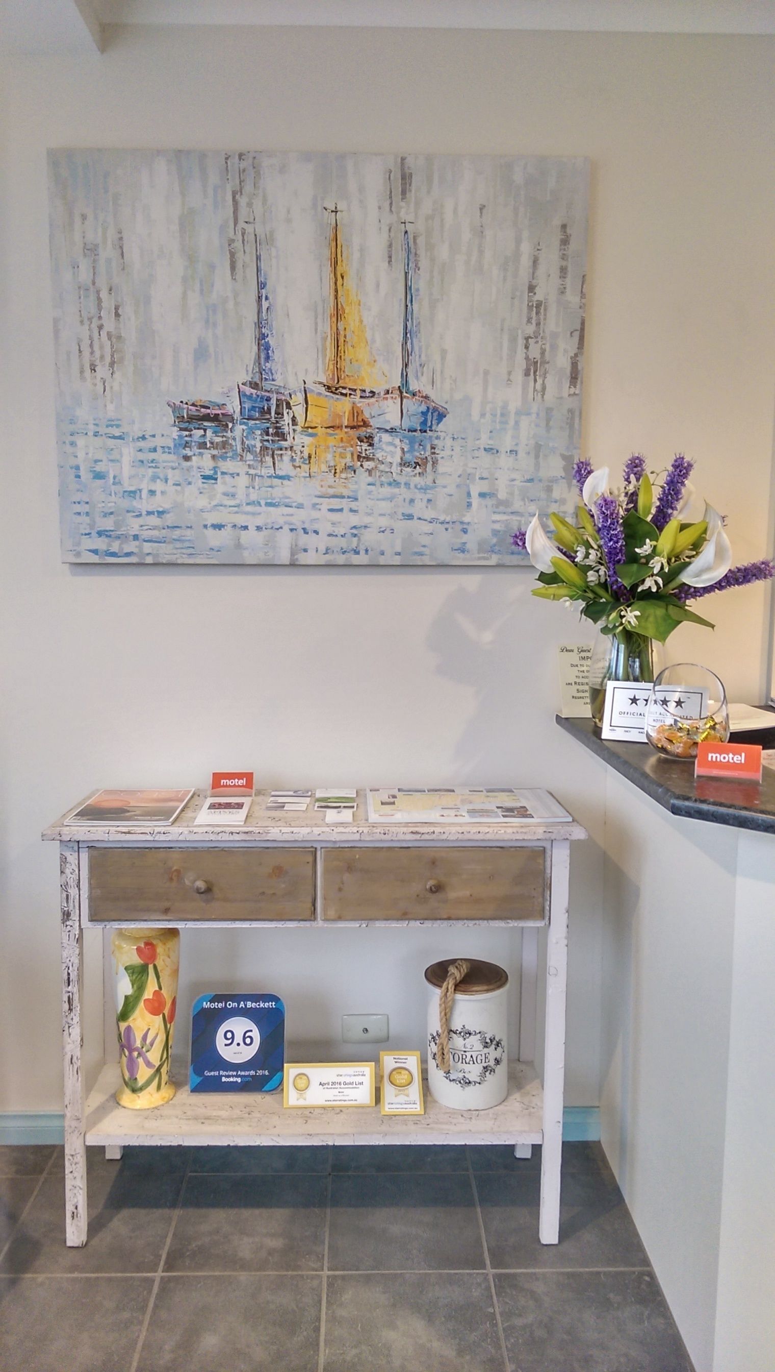 Un espace d’accueil agrémenté d’un tableau représentant des voiliers, d’un bureau avec des brochures et des trophées, et d’un vase rempli de fleurs.