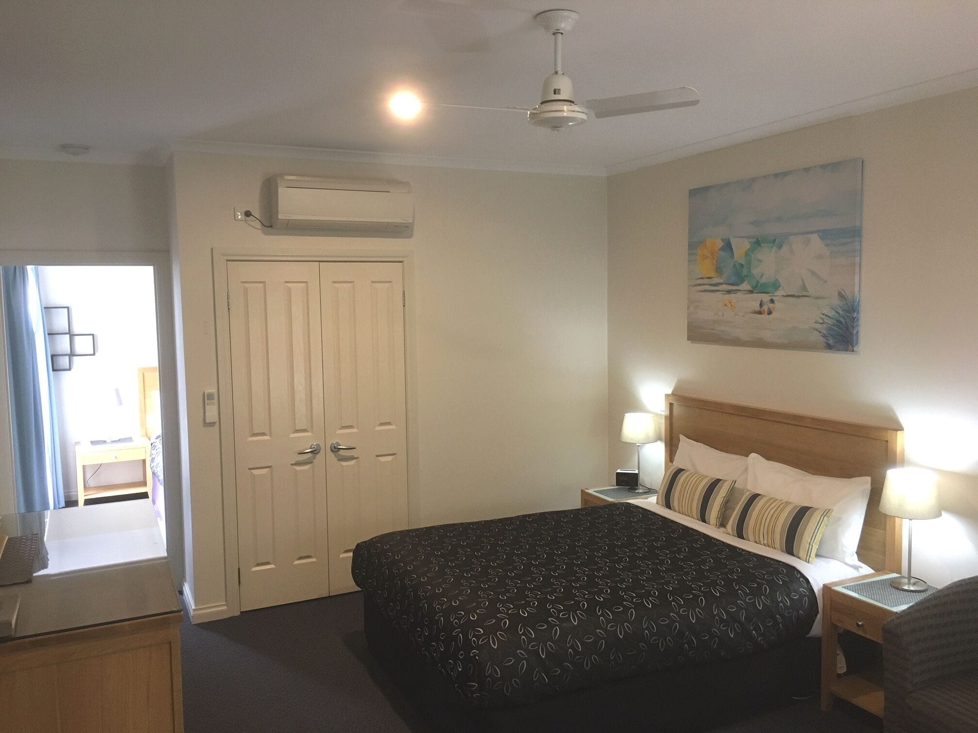 A bedroom with a bed, bedside tables, a ceiling fan, an air conditioning unit, a door, and a painting on the wall.