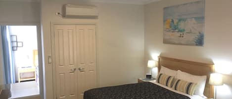 A bedroom with a bed, bedside tables, a ceiling fan, an air conditioning unit, a door, and a painting on the wall.