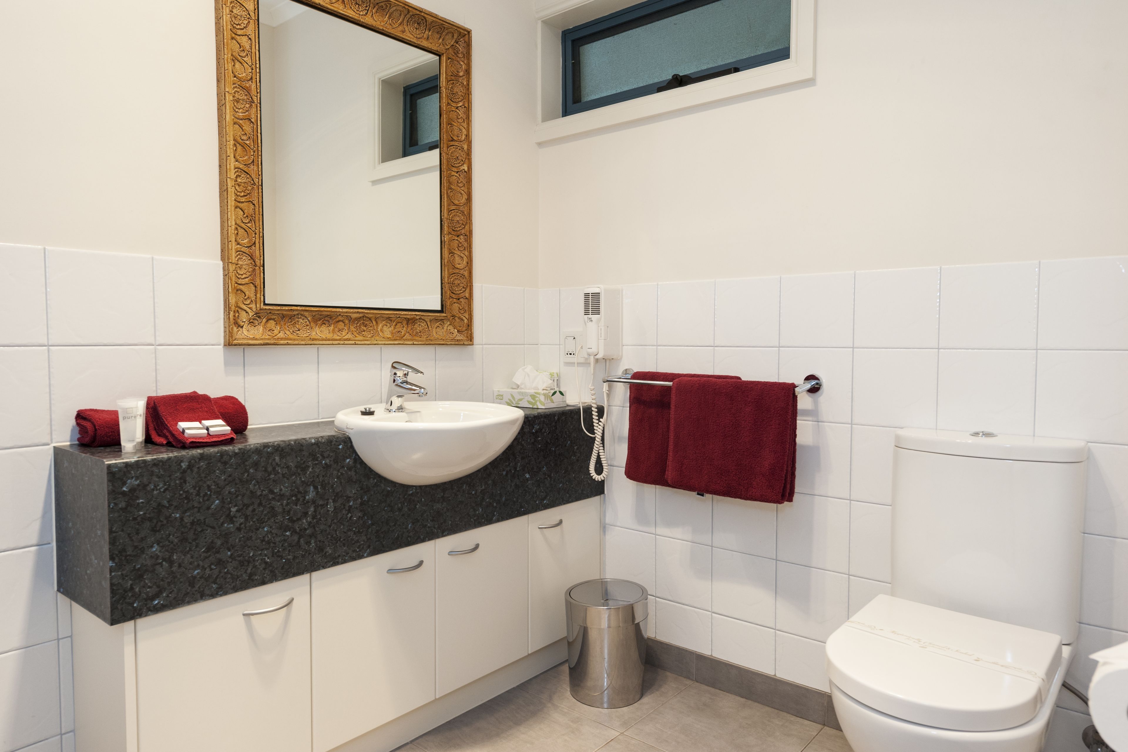 A modern bathroom with a granite countertop sink, white cabinets, a gold-framed mirror, a white toilet, and a red towel hanging on a rack.