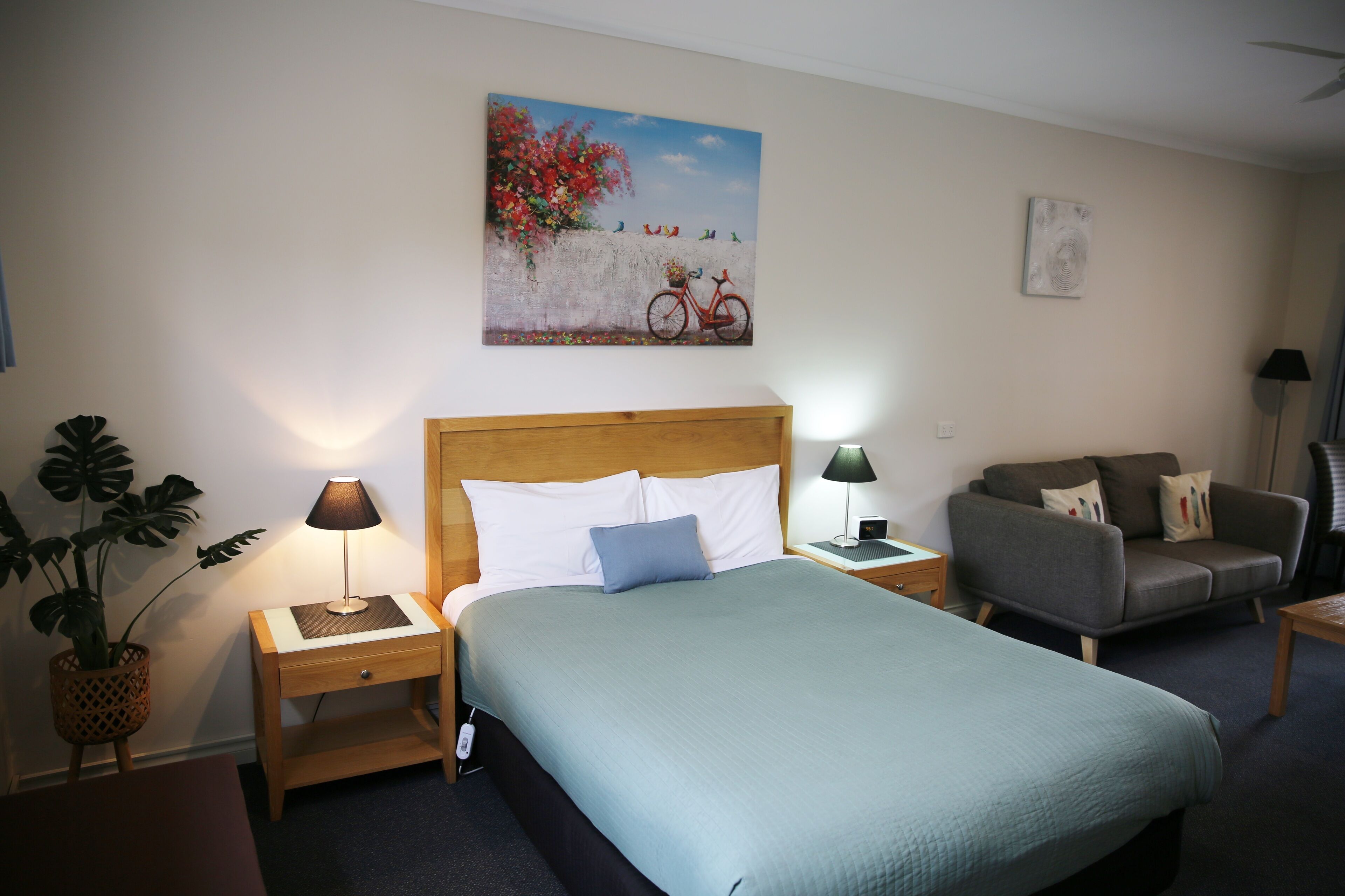 Una habitación de hotel con cama, dos mesitas de noche, lámpara, sofá, planta en maceta y un cuadro en la pared.