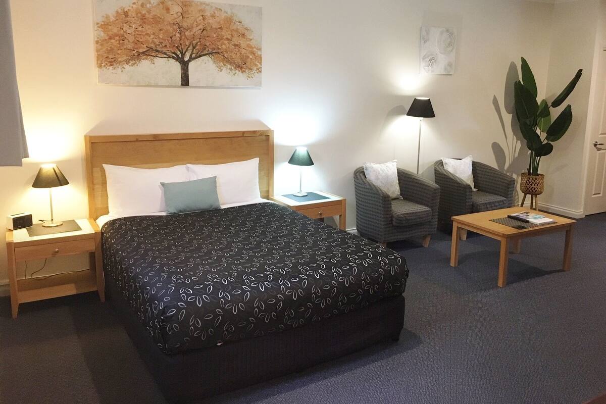 A hotel room with a large bed, two armchairs, a small table, a desk, and a potted plant.