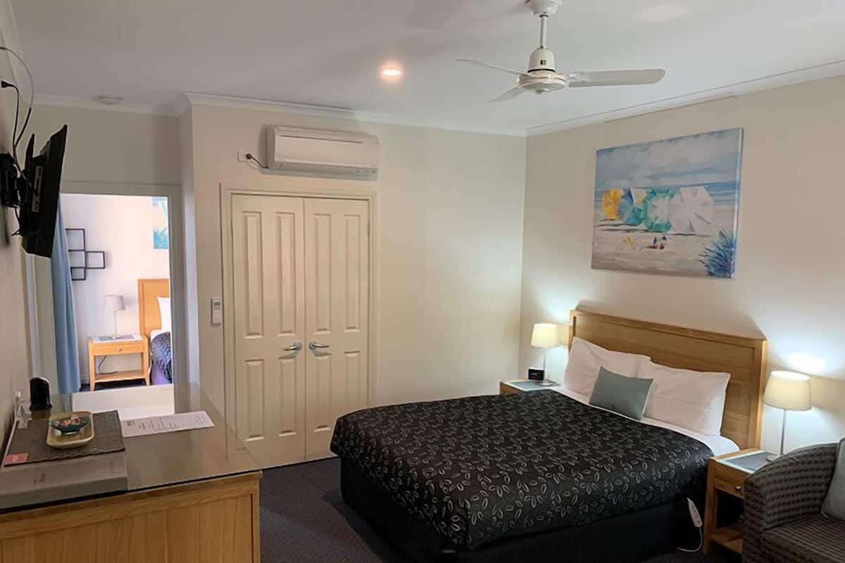 A hotel room with a bed, bedside tables, a desk, a chair, a ceiling fan, an air conditioning unit, and a painting on the wall.
