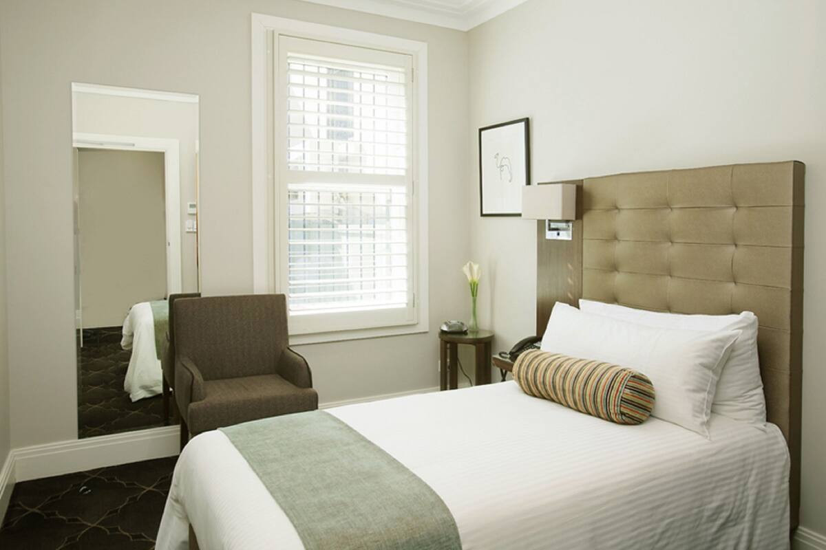 A hotel room with a large bed, a chair, a small table with a vase, and a window with blinds.