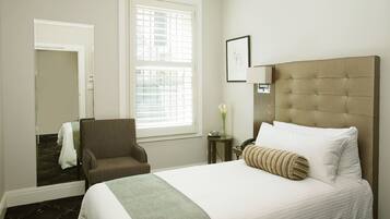 A hotel room with a large bed, a chair, a small table with a vase, and a window with blinds.