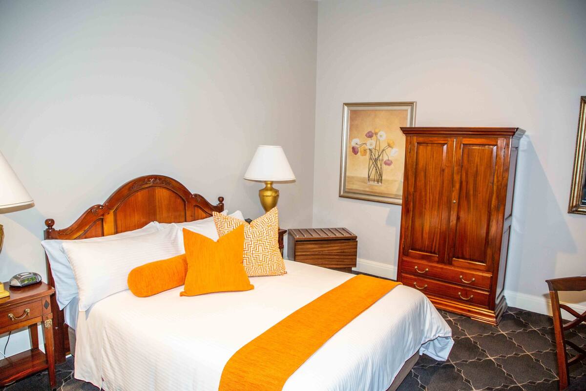 A neatly made bed with a white and orange bedspread, a wooden nightstand with a lamp, a wooden wardrobe, and a framed picture on the wall.