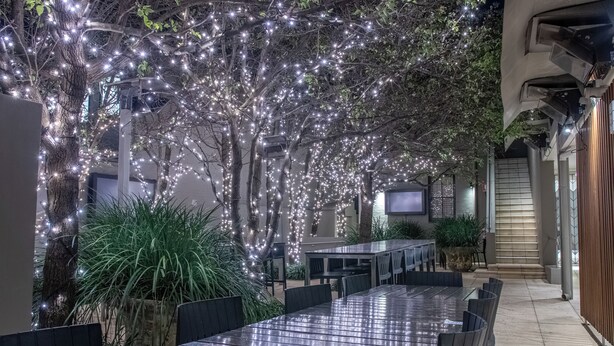 An outdoor dining area with a long table and chairs, surrounded by trees and illuminated by string lights. - Royal Hotel Randwick (Randwick)