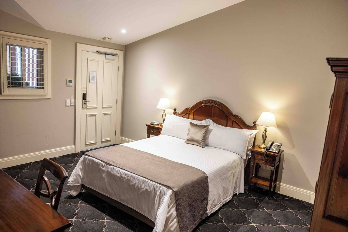A hotel room with a large bed, two bedside tables with lamps, a wooden headboard, a window with blinds, and a door.