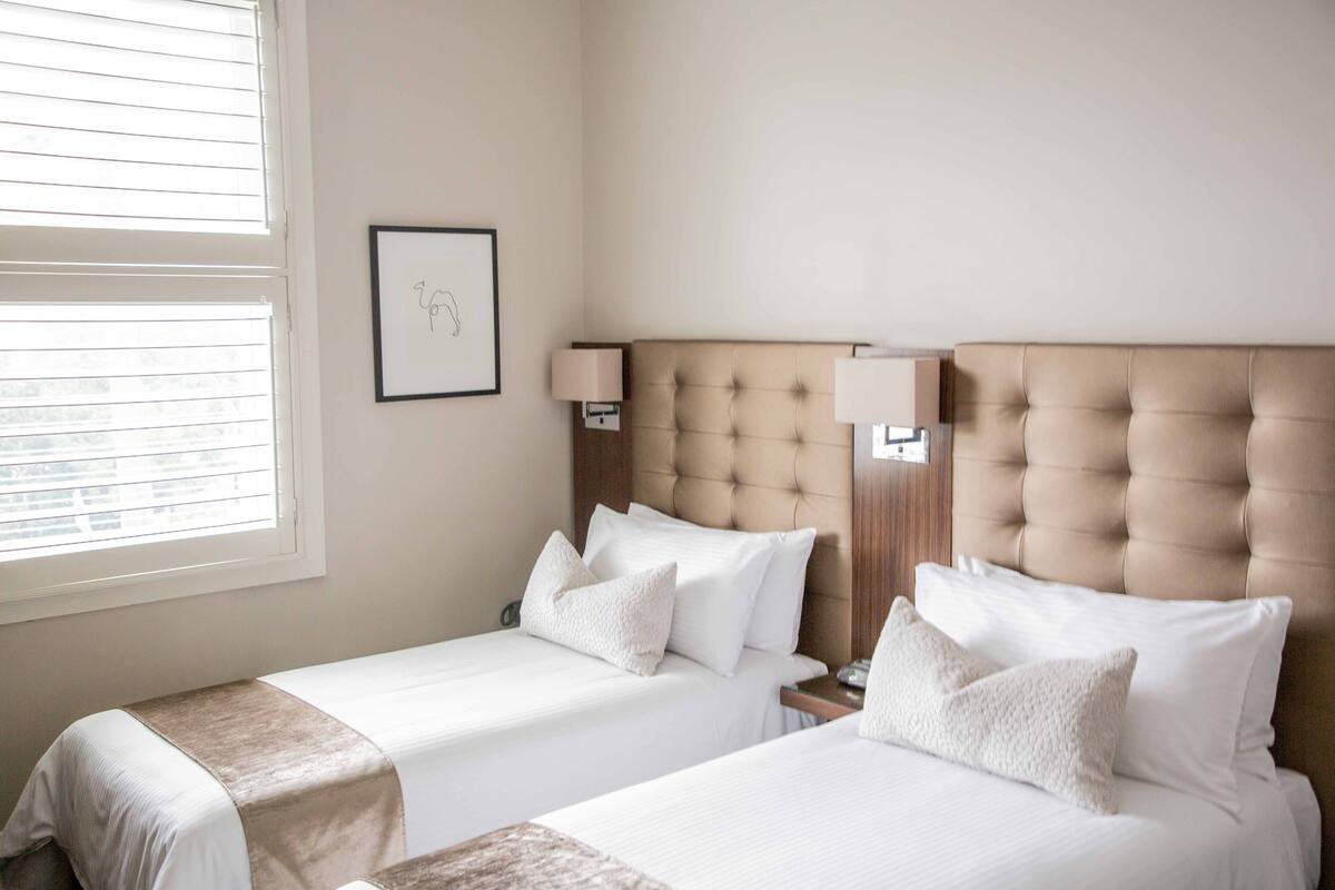 A hotel room with two beds, a window with blinds, a framed picture on the wall, and a bedside lamp.