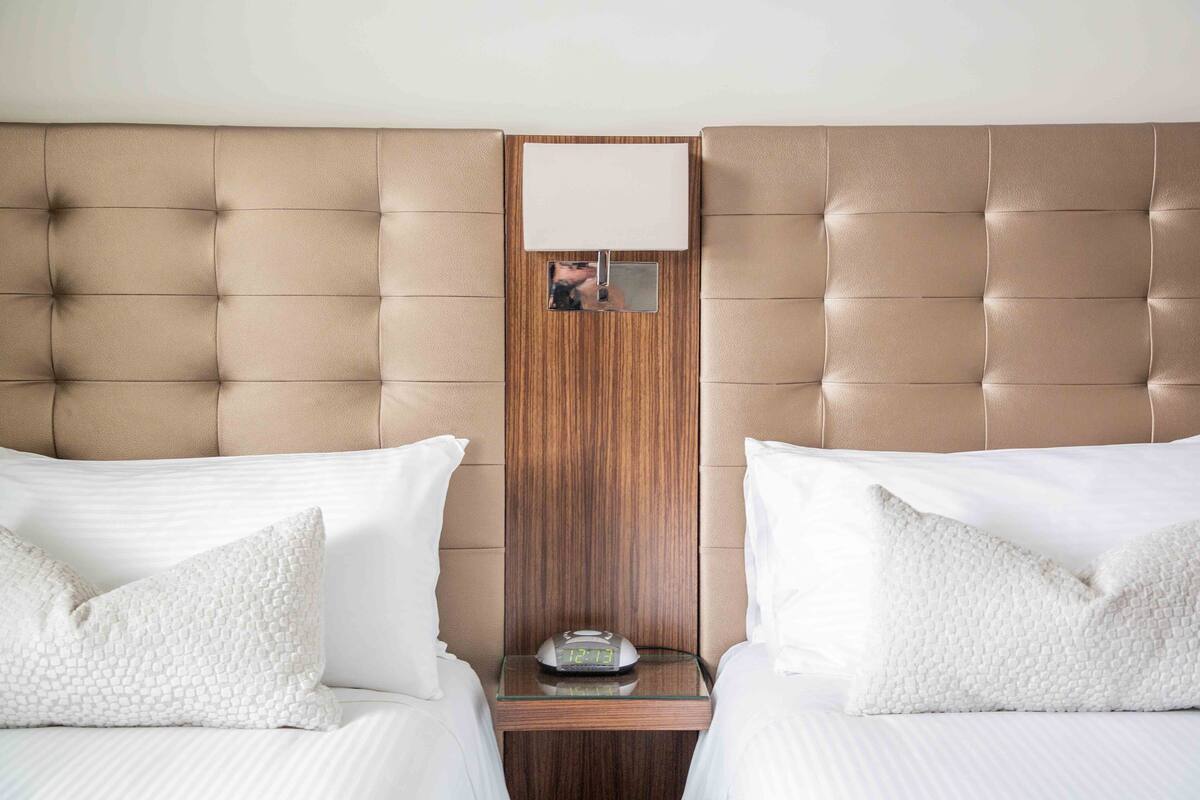 A hotel room with two beds, a wooden headboard, a bedside table with a clock, and a mirror.