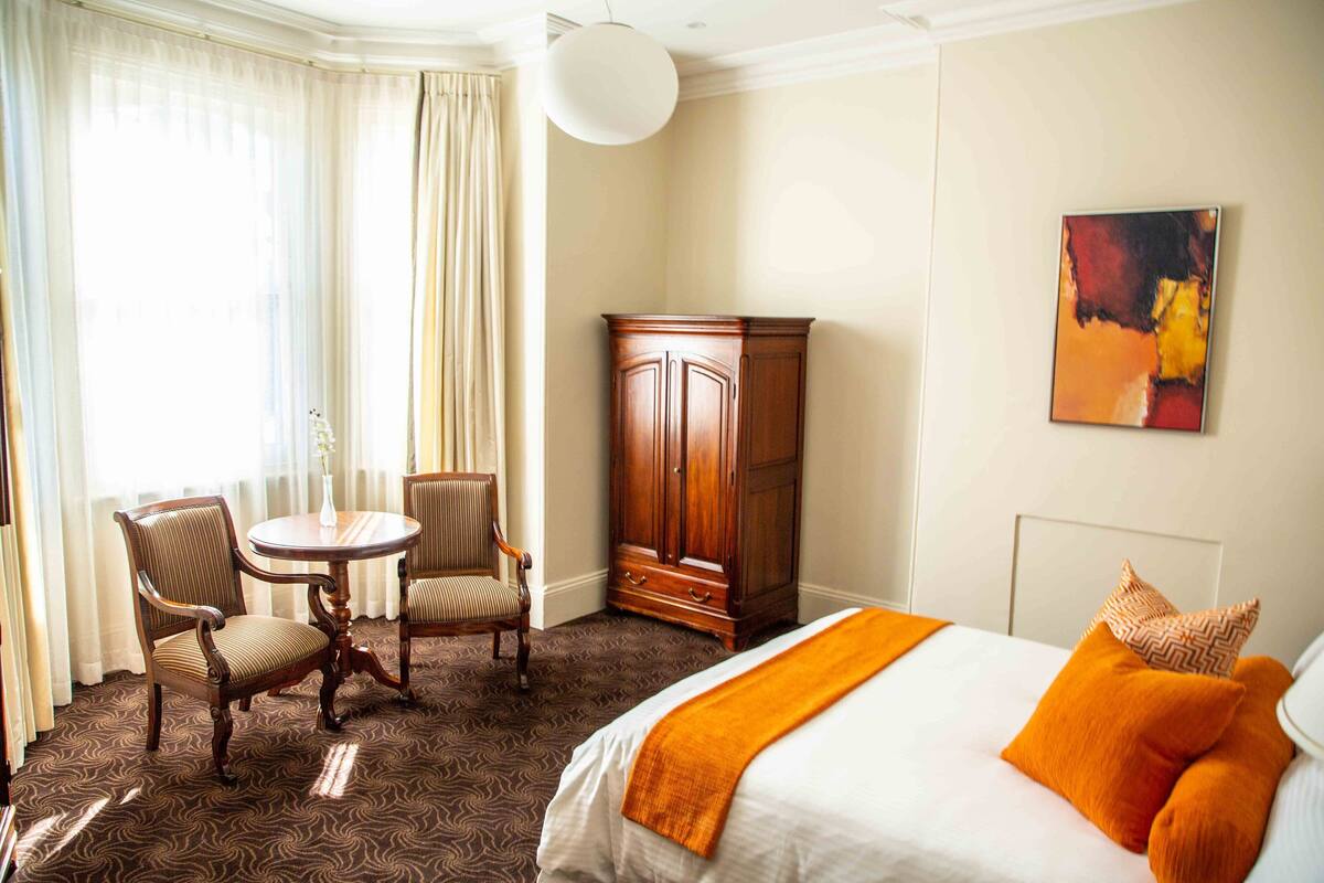 A hotel room with a bed, two chairs, a table, a wardrobe, and a painting on the wall.