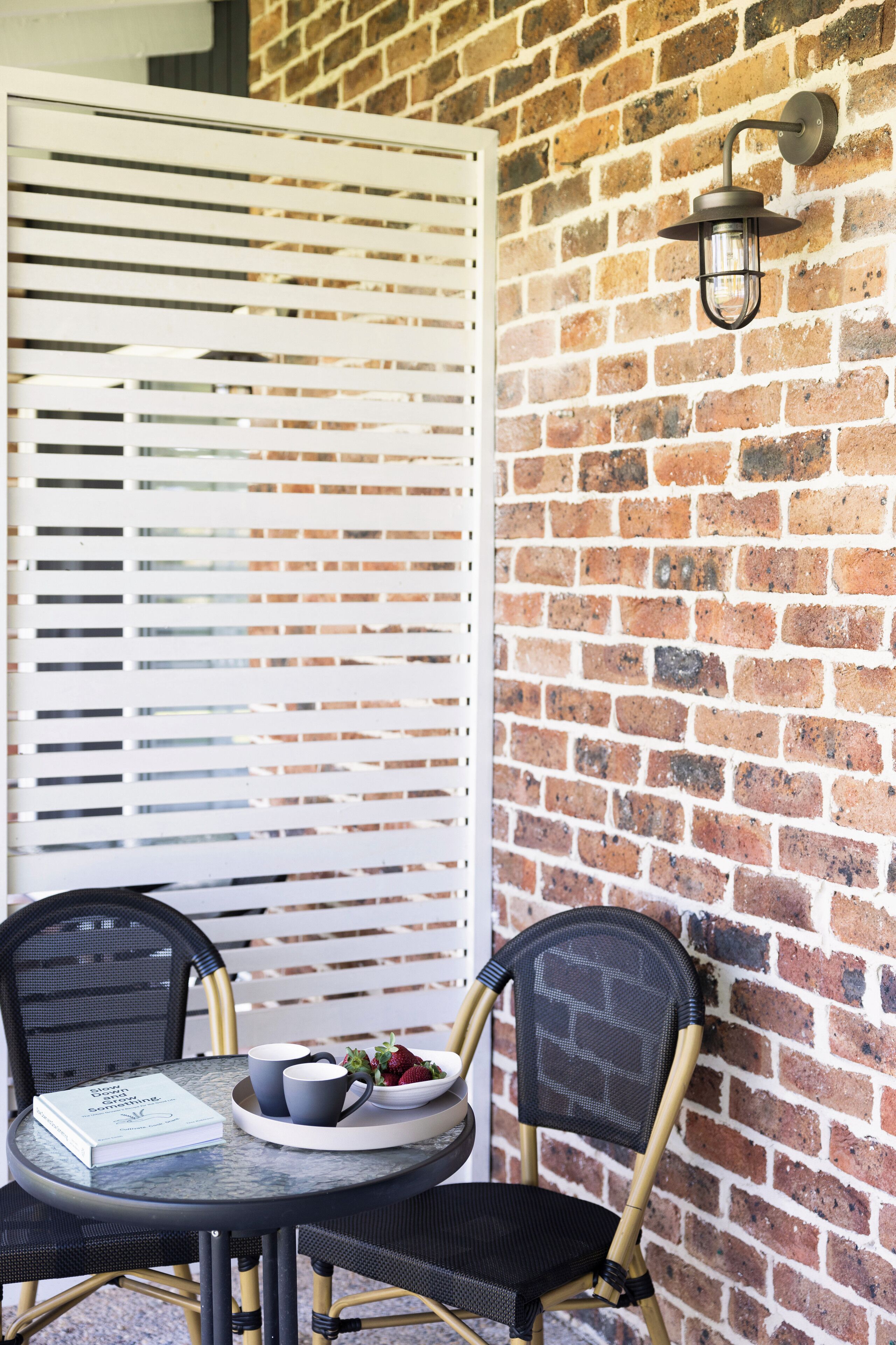 Una mesa con un libro, dos tazas y un cuenco de fruta, frente a una pared de ladrillo con un panel blanco de listones y una lámpara fijada a la pared.
