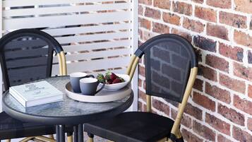 A table with a book, two cups, and a bowl of fruit, set against a brick wall with a white slatted partition and a wall-mounted light fixture.