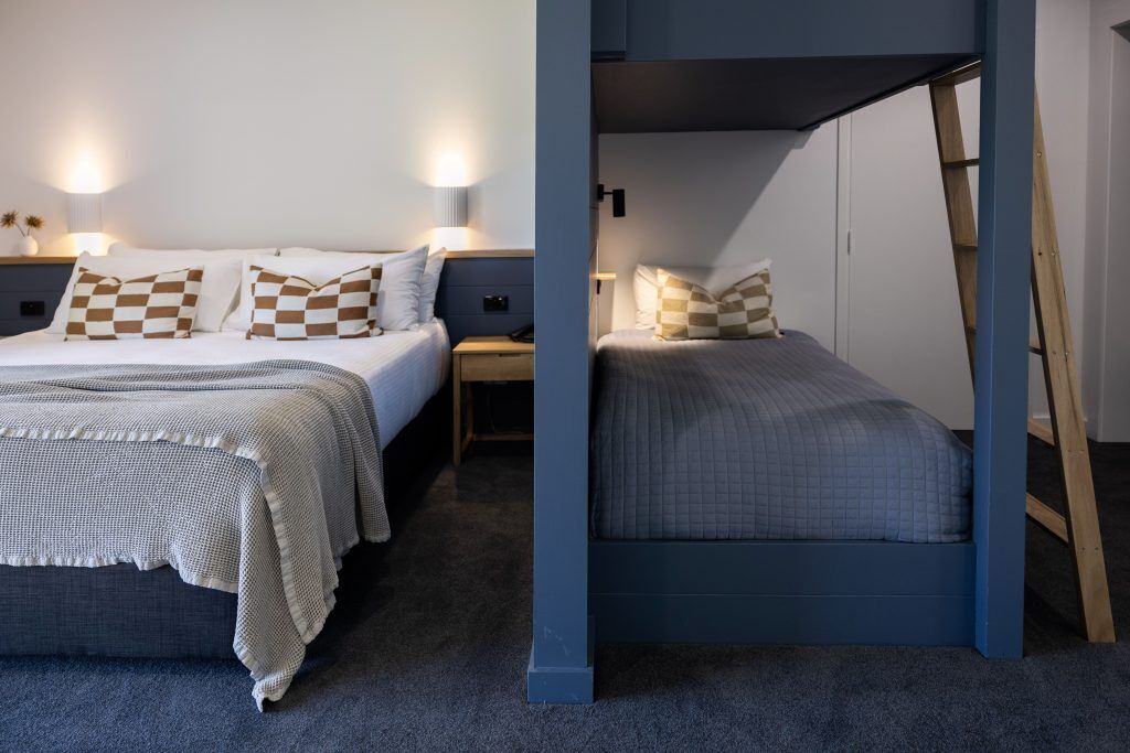 Two beds with checkered pillows, a bedside table with a lamp, and a ladder against the wall.