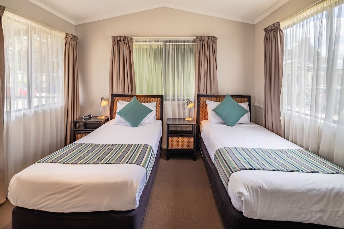 Habitación de hotel con dos camas individuales, una mesita de noche con lámpara, una ventana con cortinas traslúcidas y vista a la calle.