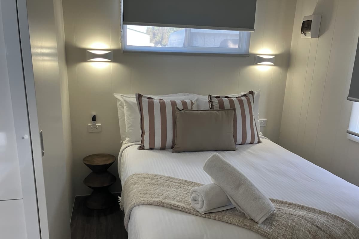 Una cama bien hecha con almohadas a rayas y una toalla enrollada, una mesita de noche y un taburete pequeño y redondo.