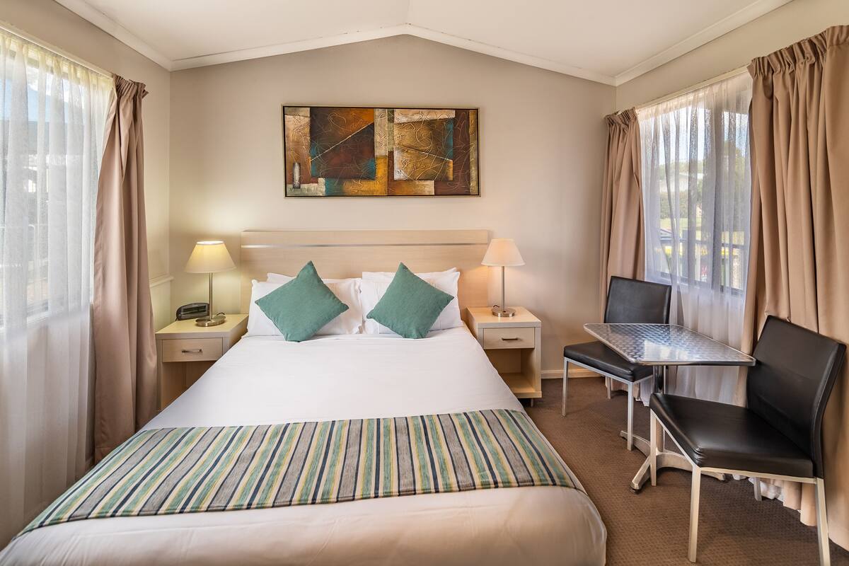 Una habitación de hotel con una cama, dos mesitas de noche con lámparas, una pequeña mesa con una silla y un cuadro en la pared.