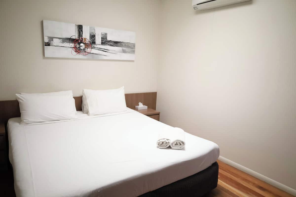 Uma cama bem arrumada com lençóis brancos, uma mesinha de cabeceira com caixa de lenços e um quadro abstrato pendurado na parede.