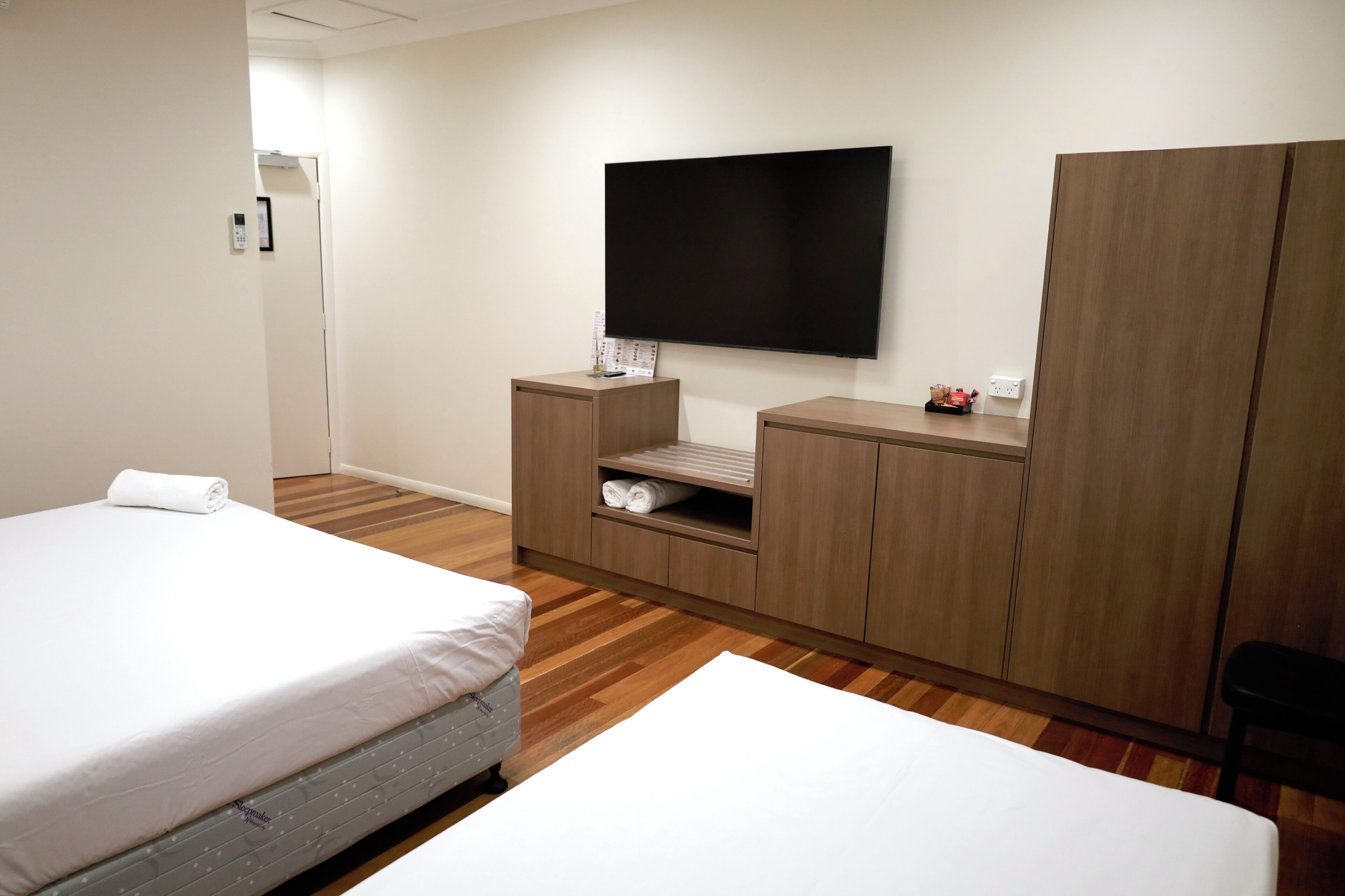 Habitación de hotel con dos camas, televisión montada en la pared, muebles de madera y un estante pequeño con toallas.