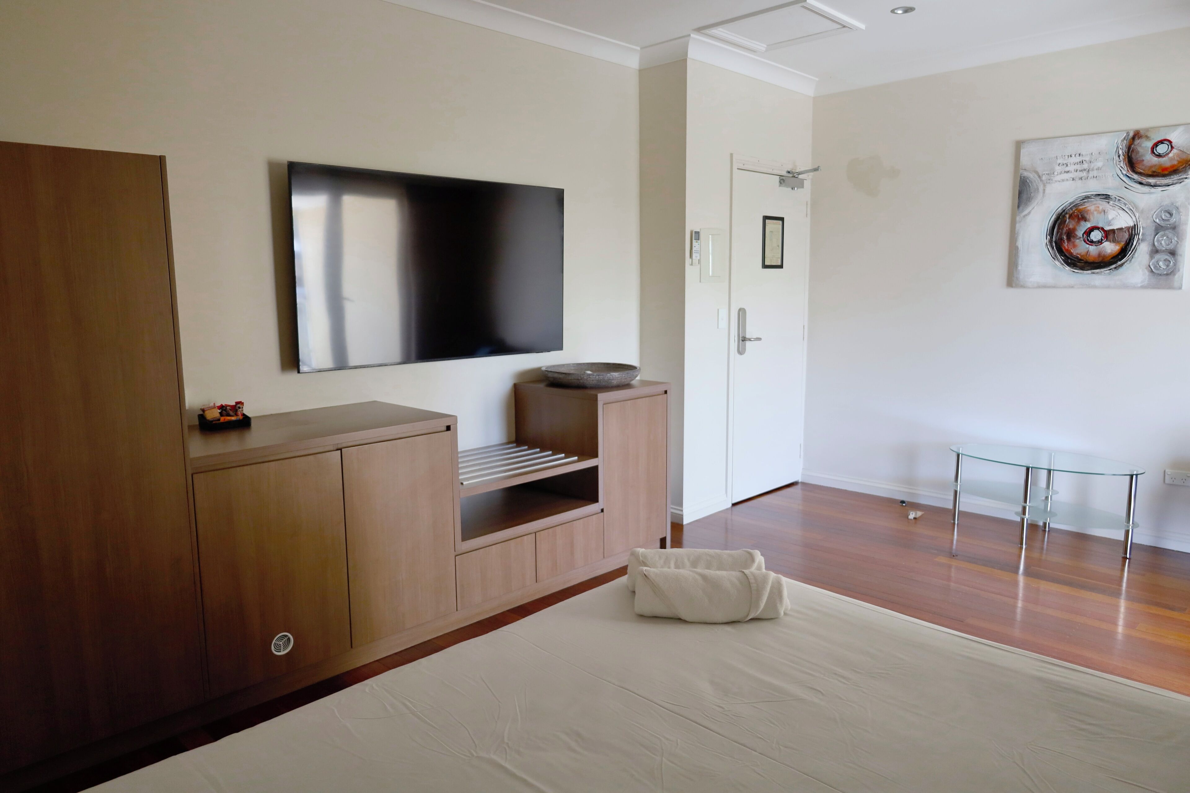 A modern hotel room with a large flat-screen TV mounted on the wall, a wooden cabinet with built-in shelves, a glass coffee table, and a large bed with a white comforter.