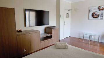 A modern hotel room with a large flat-screen TV mounted on the wall, a wooden cabinet with built-in shelves, a glass coffee table, and a large bed with a white comforter.