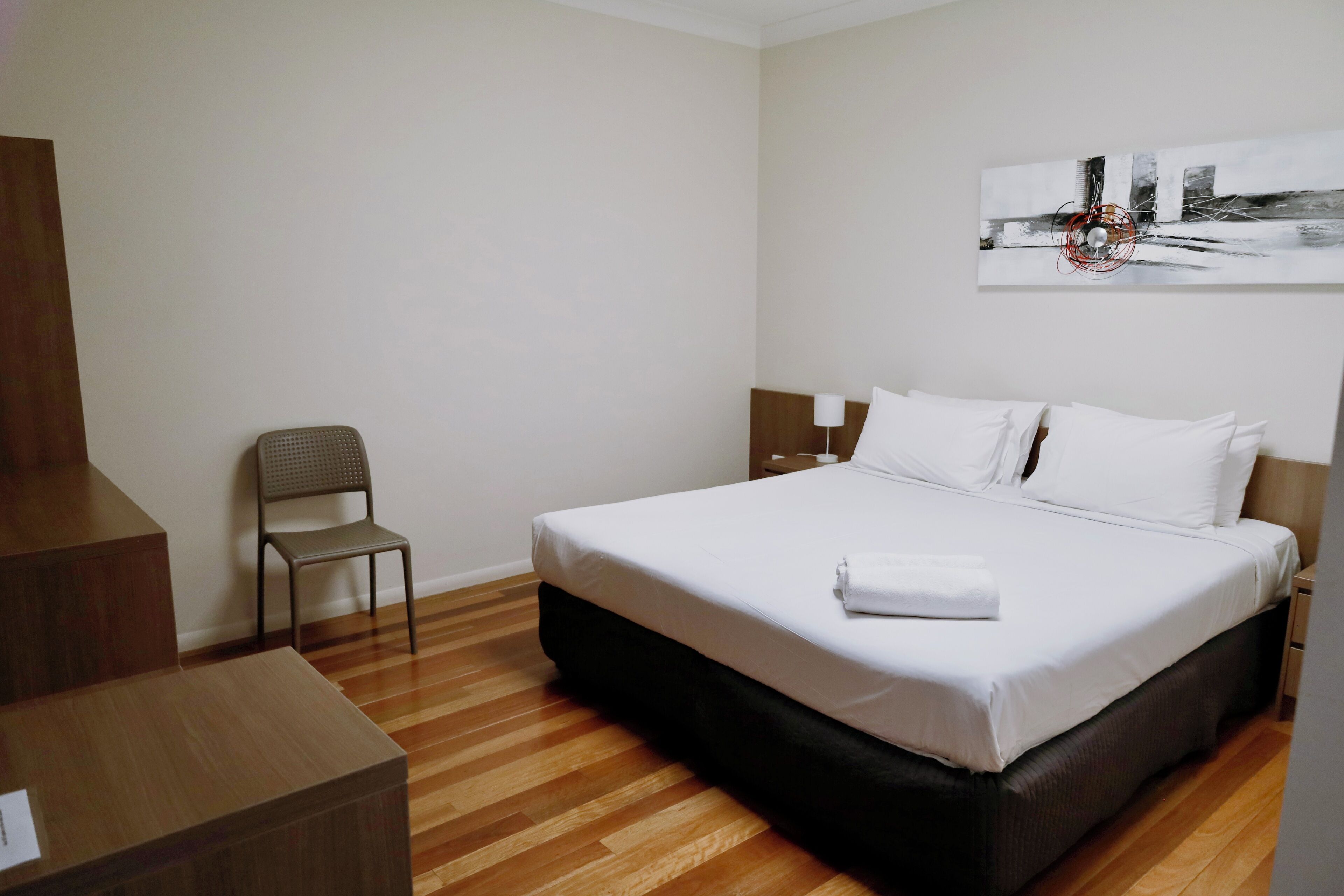 A hotel room with a bed, a chair, a desk, and a painting on the wall.