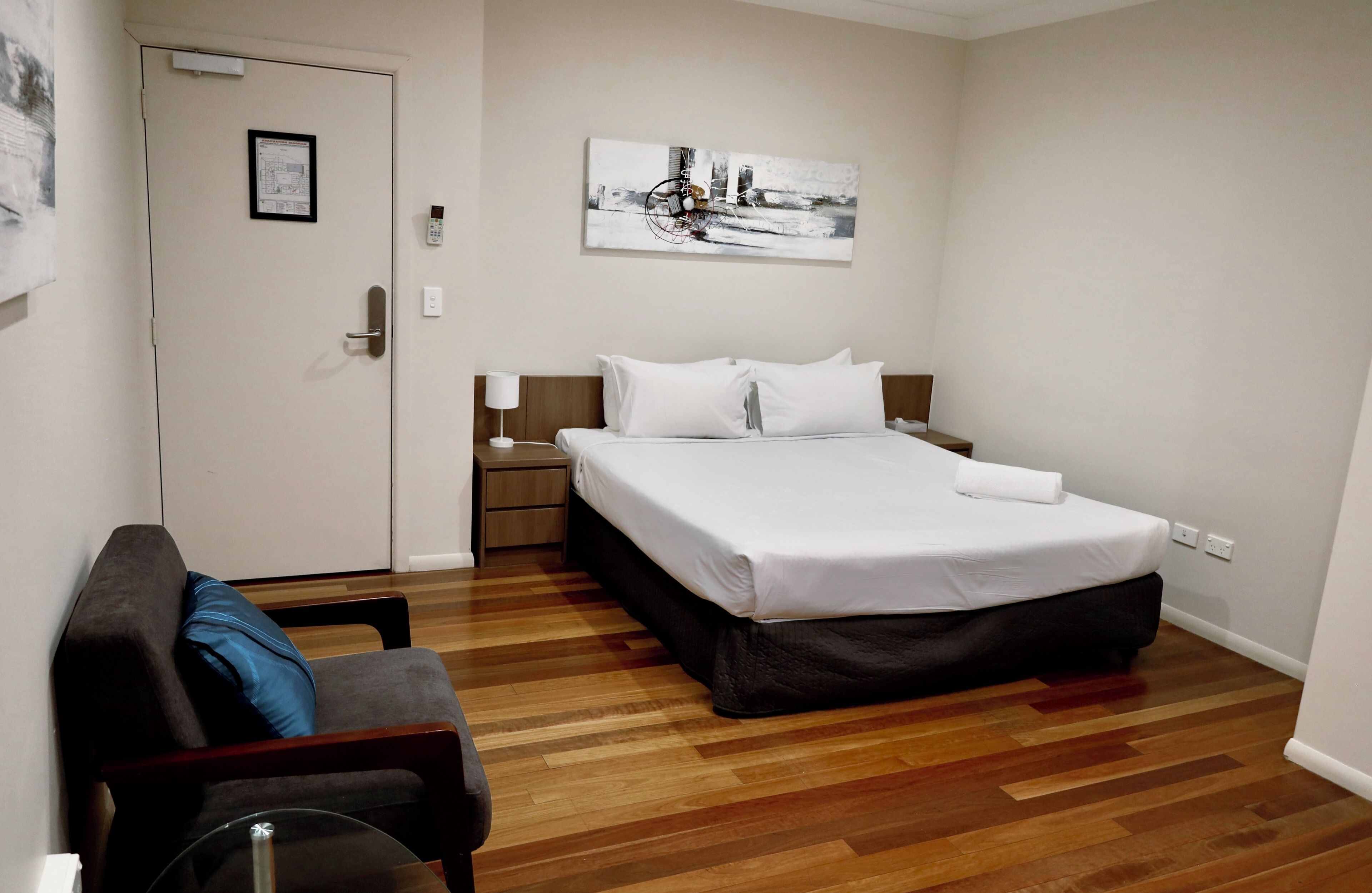 Een hotelkamer met een bed, nachtkastje, lamp, stoel en een houten vloer.