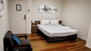 A hotel room with a bed, a nightstand, a lamp, a chair, and a wooden floor.