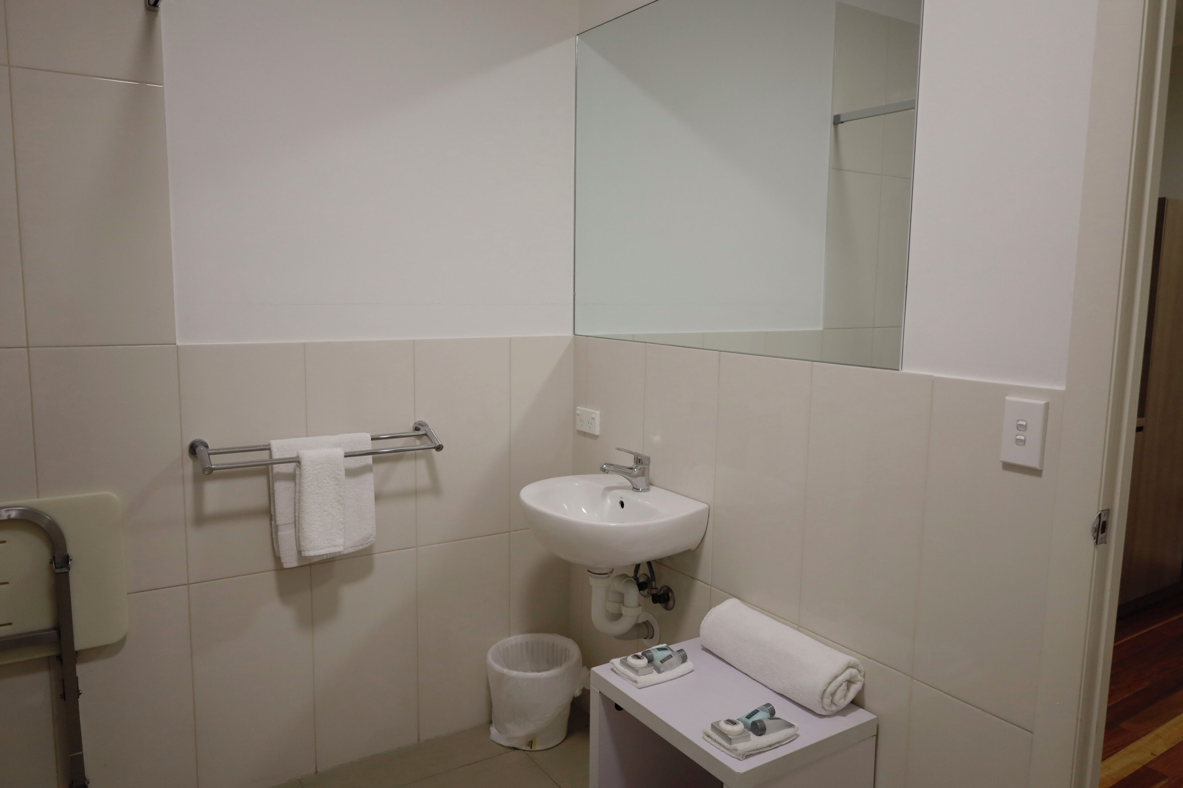 A bathroom with white tiles, a sink, a mirror, a towel rack, a trash bin, and a small table with toiletries.