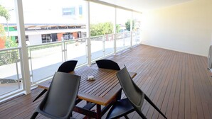 Ein Balkon mit Holztisch und -stühlen, großen Fenstern und Blick auf ein Gebäude und Bäume.