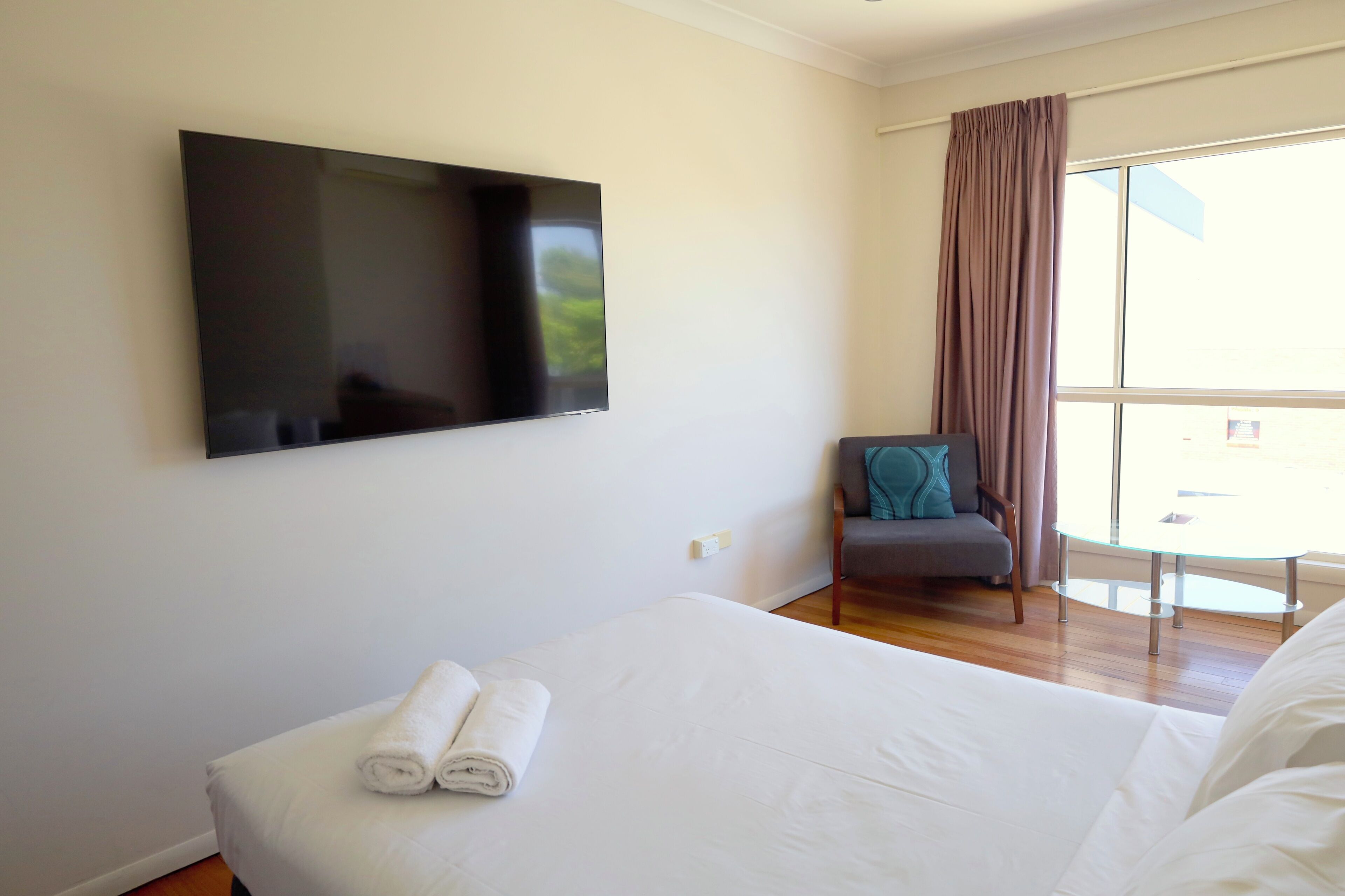 A hotel room with a bed, a flat-screen TV mounted on the wall, a chair, and a window with curtains.