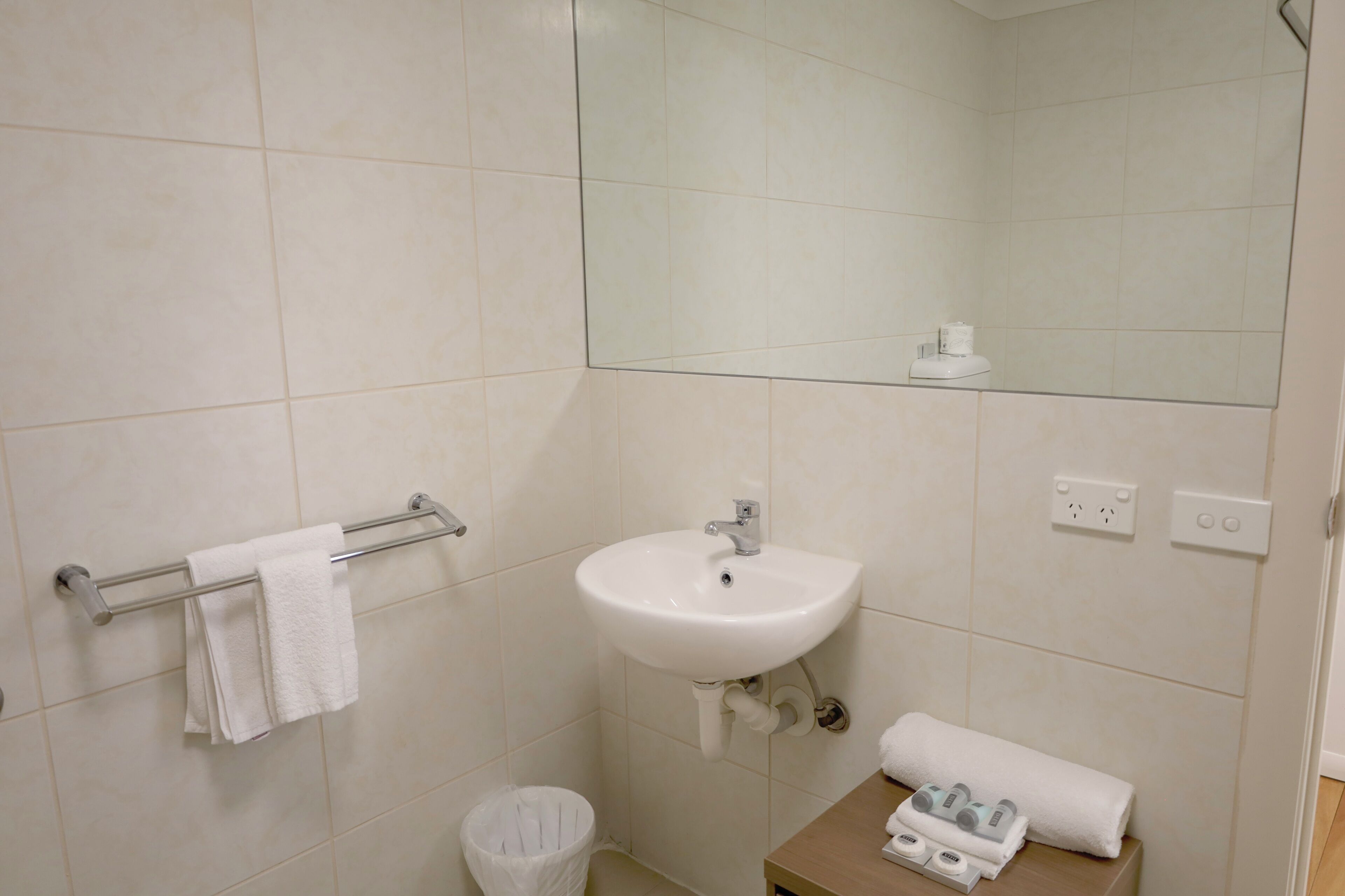 Un baño con lavabo, espejo, toallero y una pequeña mesa con artículos de aseo.