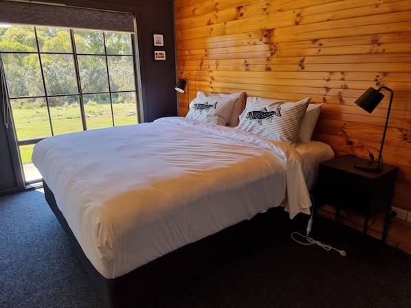 Un dormitorio con una cama grande, paredes de madera, una ventana con vistas a follaje y una mesita de noche con una lámpara.