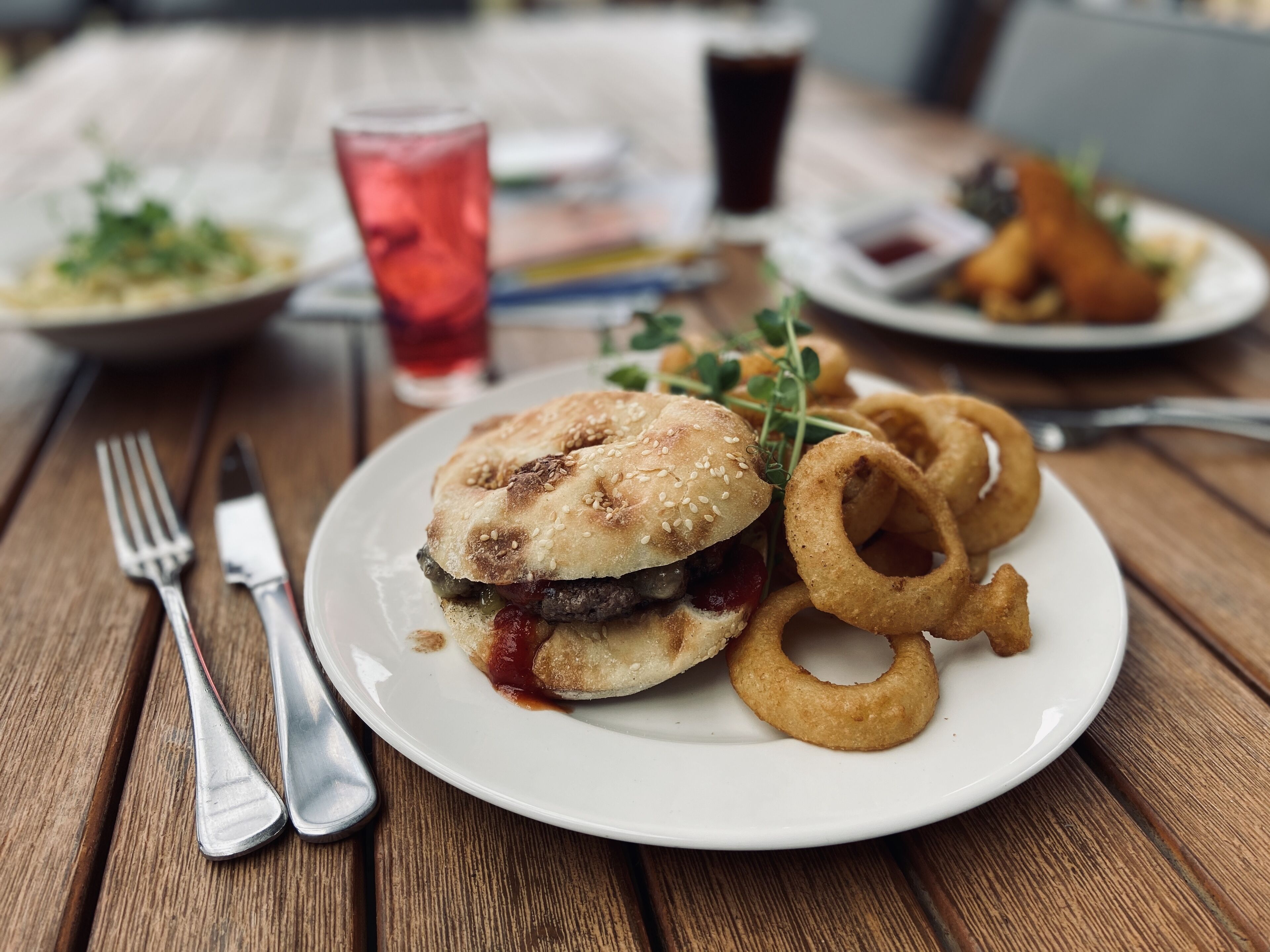 Ein Gericht bestehend aus einem Burger mit Sesam, Zwiebelringen und einem Glas rotem Getränk auf einem Holztisch.