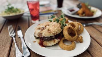 Ein Gericht bestehend aus einem Burger mit Sesam, Zwiebelringen und einem Glas rotem Getränk auf einem Holztisch.