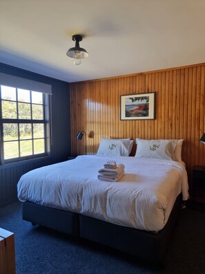 A bedroom with a wooden paneled wall, a large bed with white linens, a framed landscape painting, and a ceiling fan. - Great Lake Hotel (Miena)