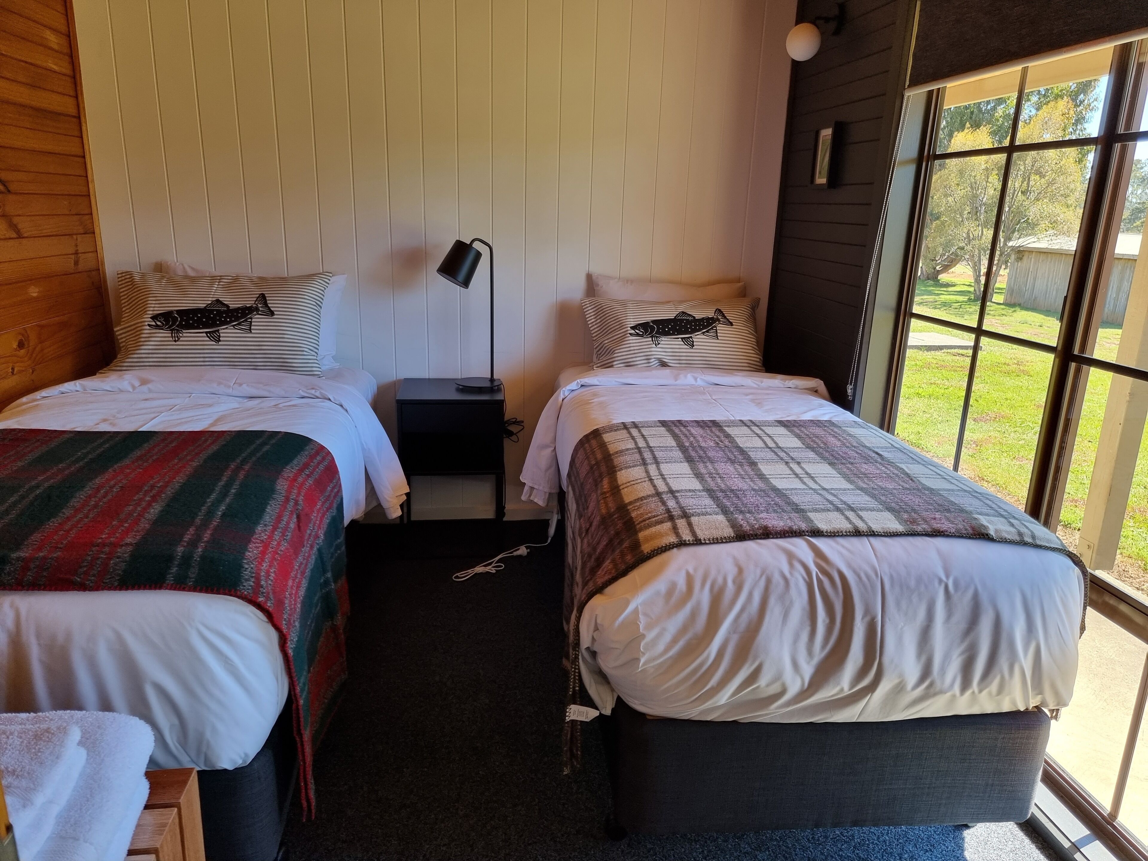 A room with two beds, each with a plaid blanket and a pillow with a fish design. There is a nightstand with a lamp between the beds.