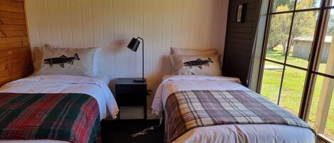 A room with two beds, each with a plaid blanket and a pillow with a fish design. There is a nightstand with a lamp between the beds.