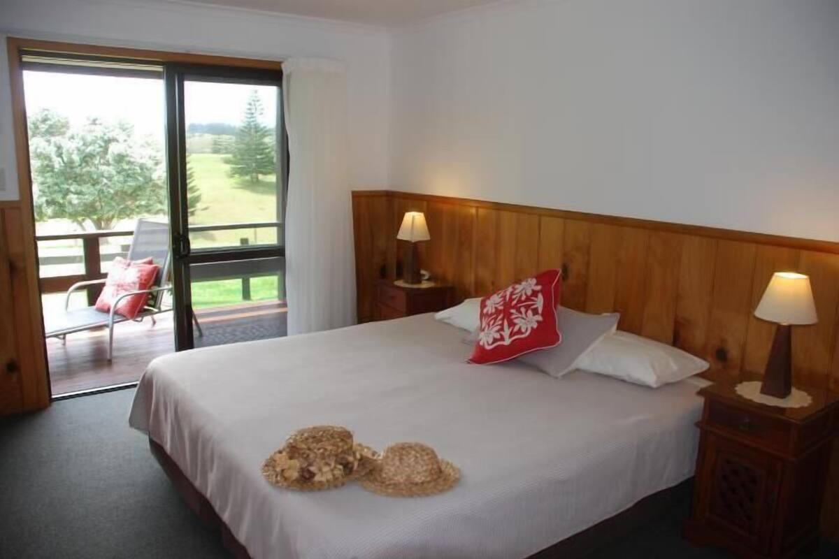 Quarto com uma cama grande, duas luminárias de cabeceira, uma cabeceira de madeira, um travesseiro vermelho e vista para um deck e árvores através de uma porta de correr de vidro.