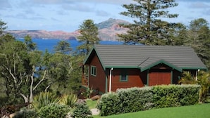 Une petite maison à un seul étage, avec un toit rouge et des bordures vertes, entourée d’une végétation luxuriante et offrant une vue sur la mer.