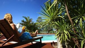 Une personne se détend sur une chaise longue en bois, près d’une piscine, entourée de plantes tropicales.