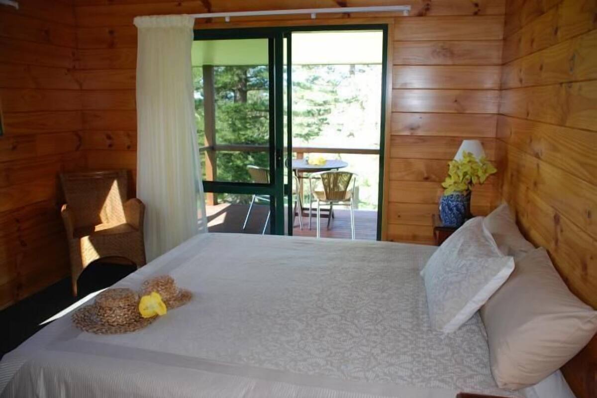 Quarto de cabana de madeira com uma cama, uma cadeira, uma pequena mesa e vista para uma varanda com mobiliário exterior.