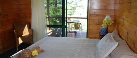 A wooden cabin room with a bed, a chair, a small table, and a view of a balcony with outdoor furniture.