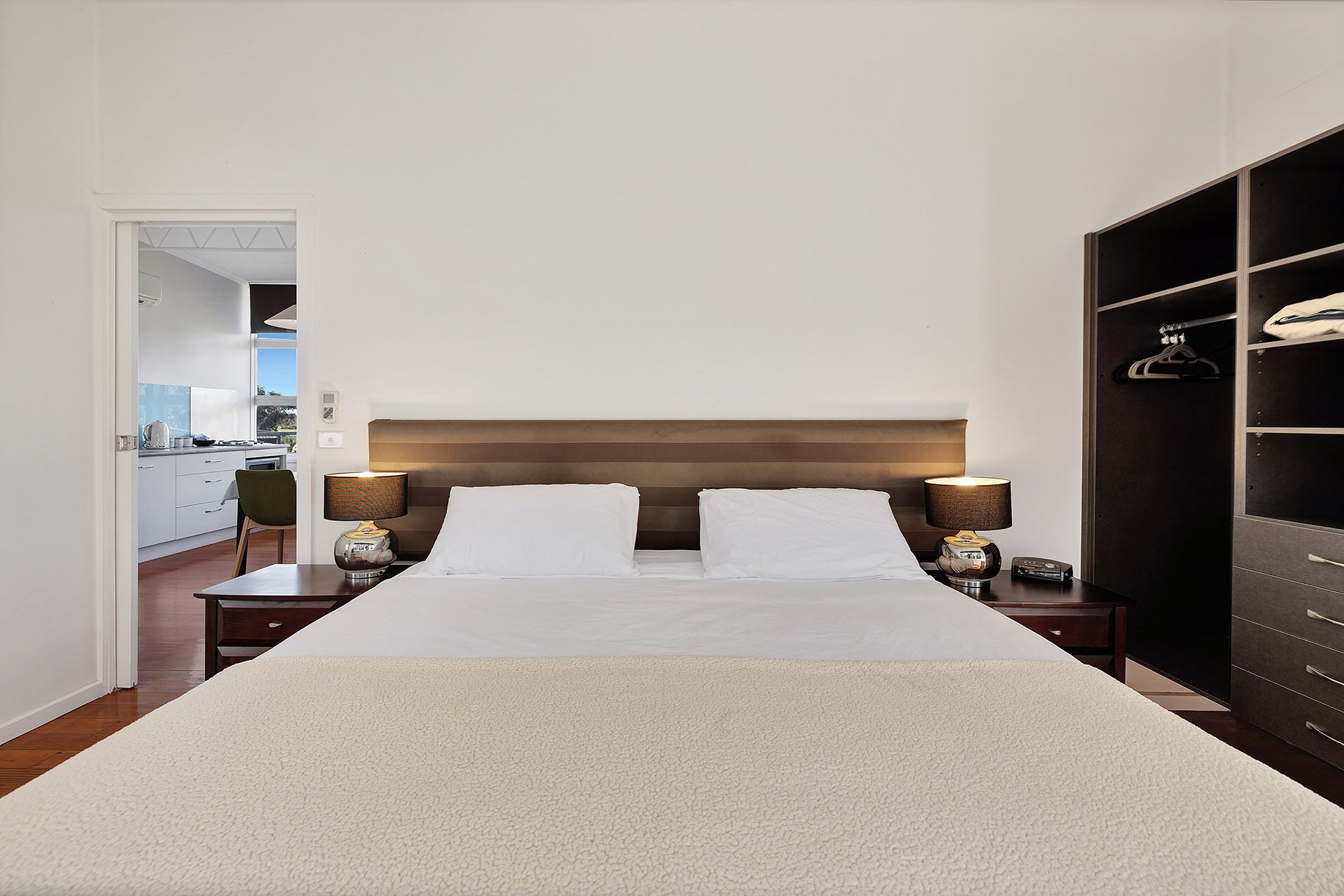 A hotel room with a large bed, two bedside tables with lamps, a wardrobe, and a view of the kitchen.
