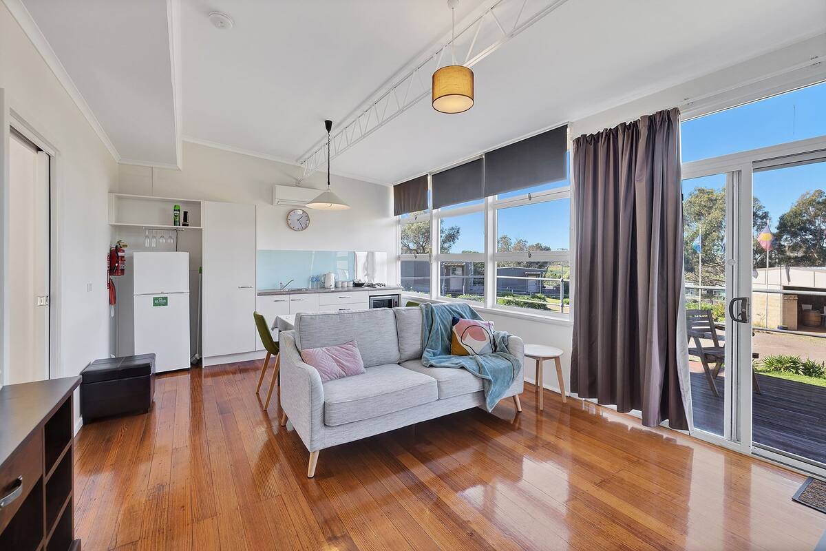 A modern living room with a grey sofa, wooden flooring, a kitchen area with a fridge, and large windows with curtains.