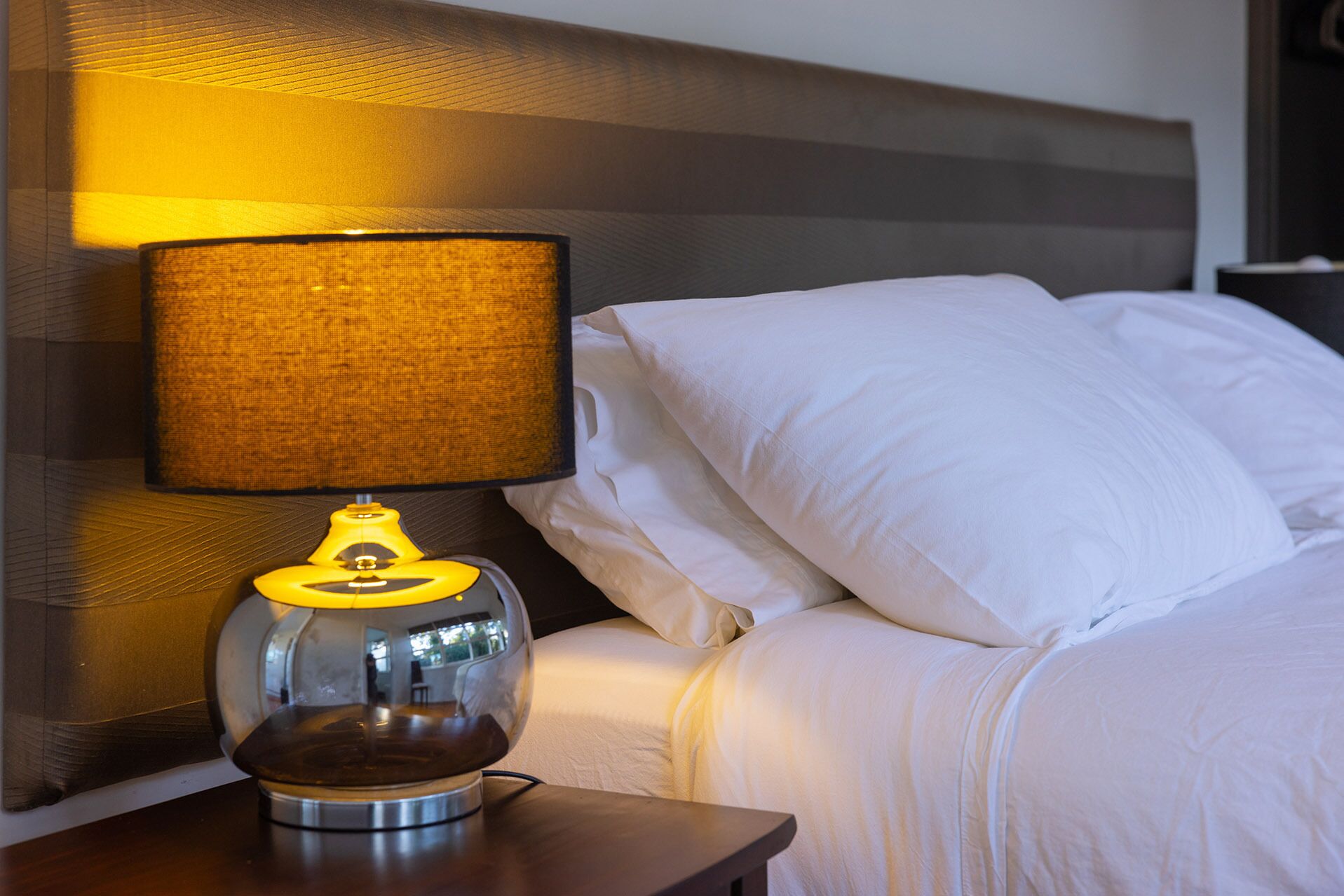 A bed with white linens and a lamp with a yellow shade on a wooden nightstand.