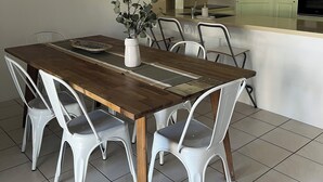 Un coin repas avec une table en bois, des chaises blanches, un vase avec une plante, et une cuisine en arrière-plan.