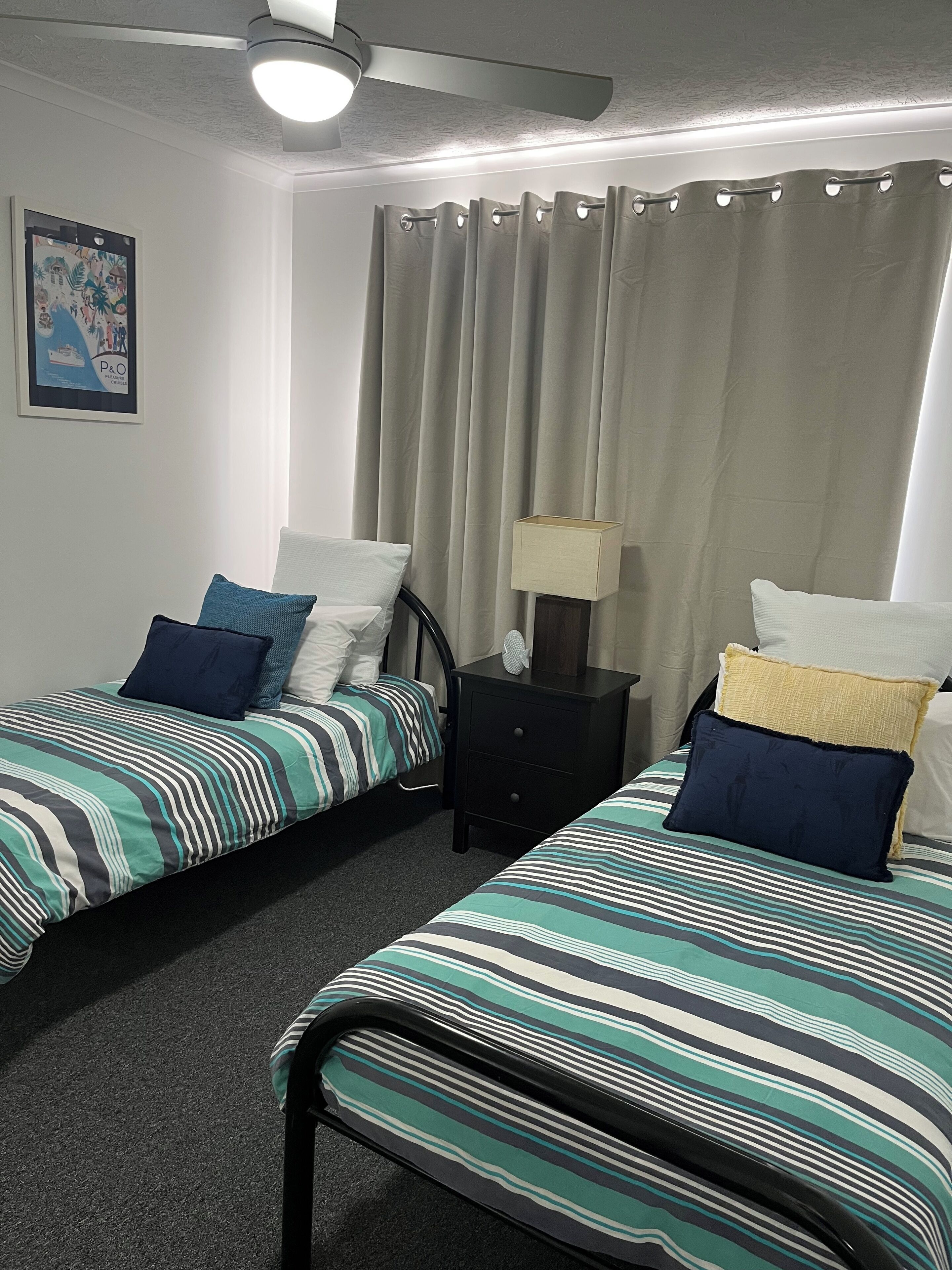 A room with two single beds, a ceiling fan, a framed picture on the wall, and a nightstand with a lamp.