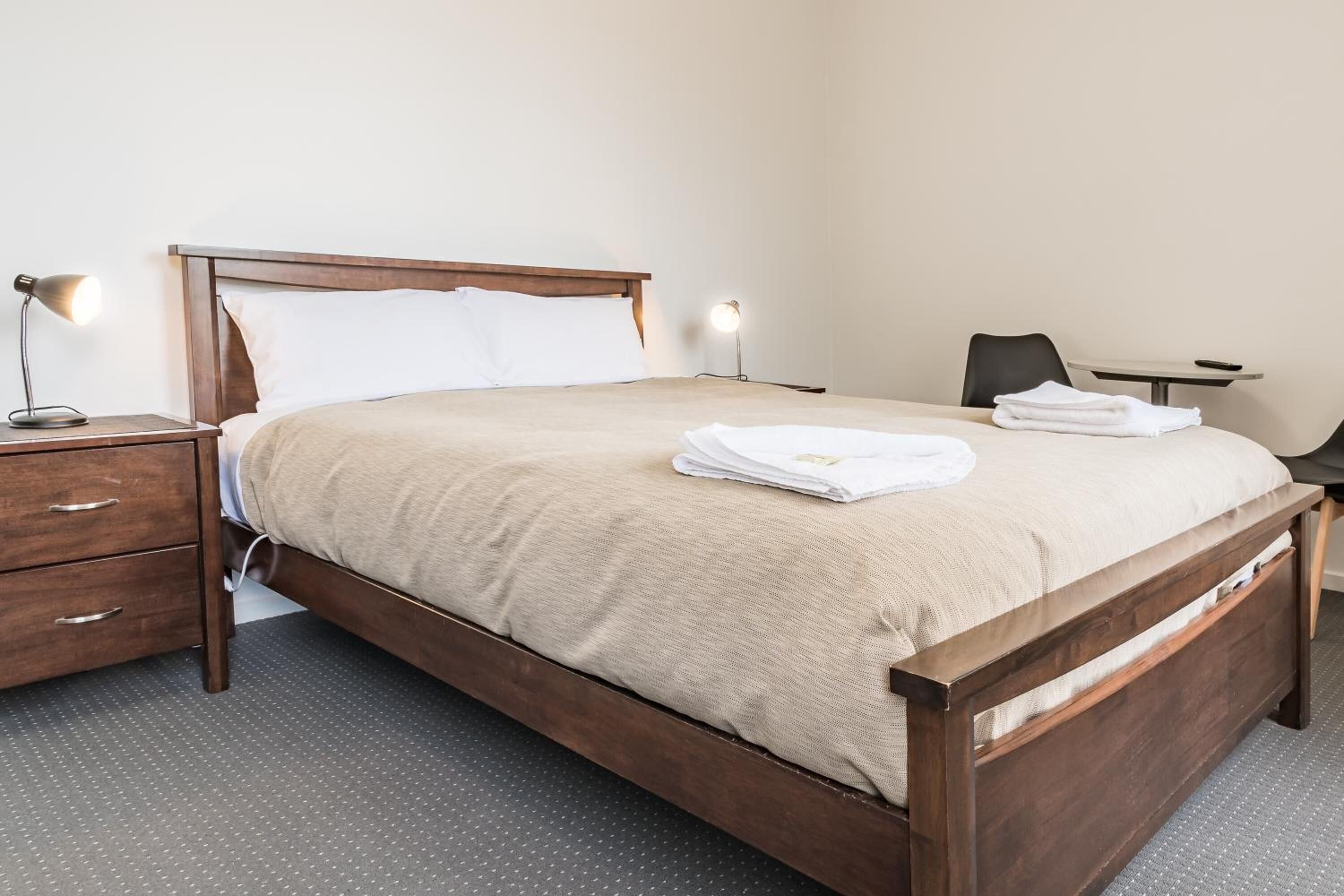 A neatly made bed with a wooden frame, a bedside table with a lamp, and a chair in a simple room with a gray carpet.